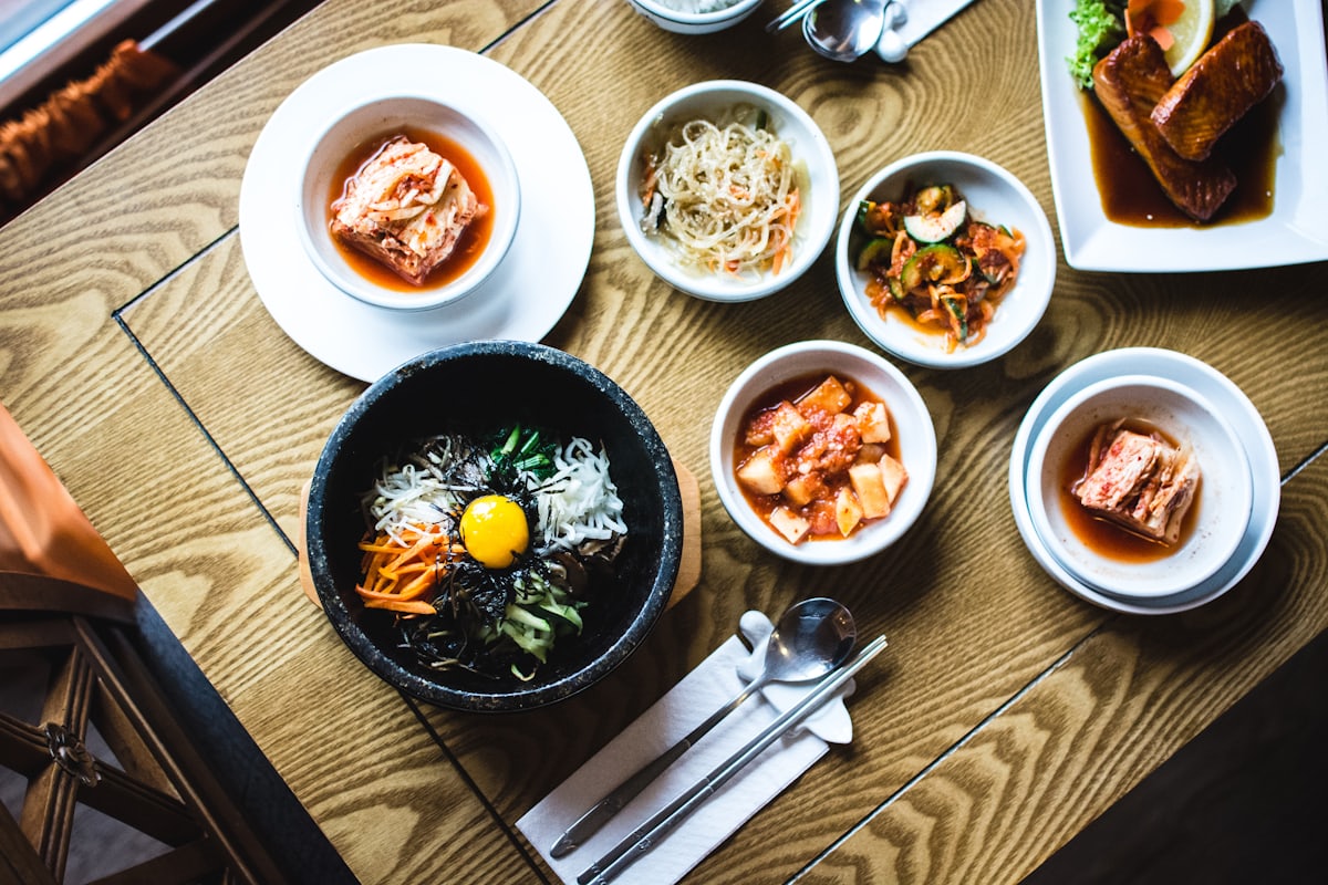 La table vue du dessus : bols de kimchi, de pousses de soja et bibimbap dans un bol noir, serrés sur le bois clair