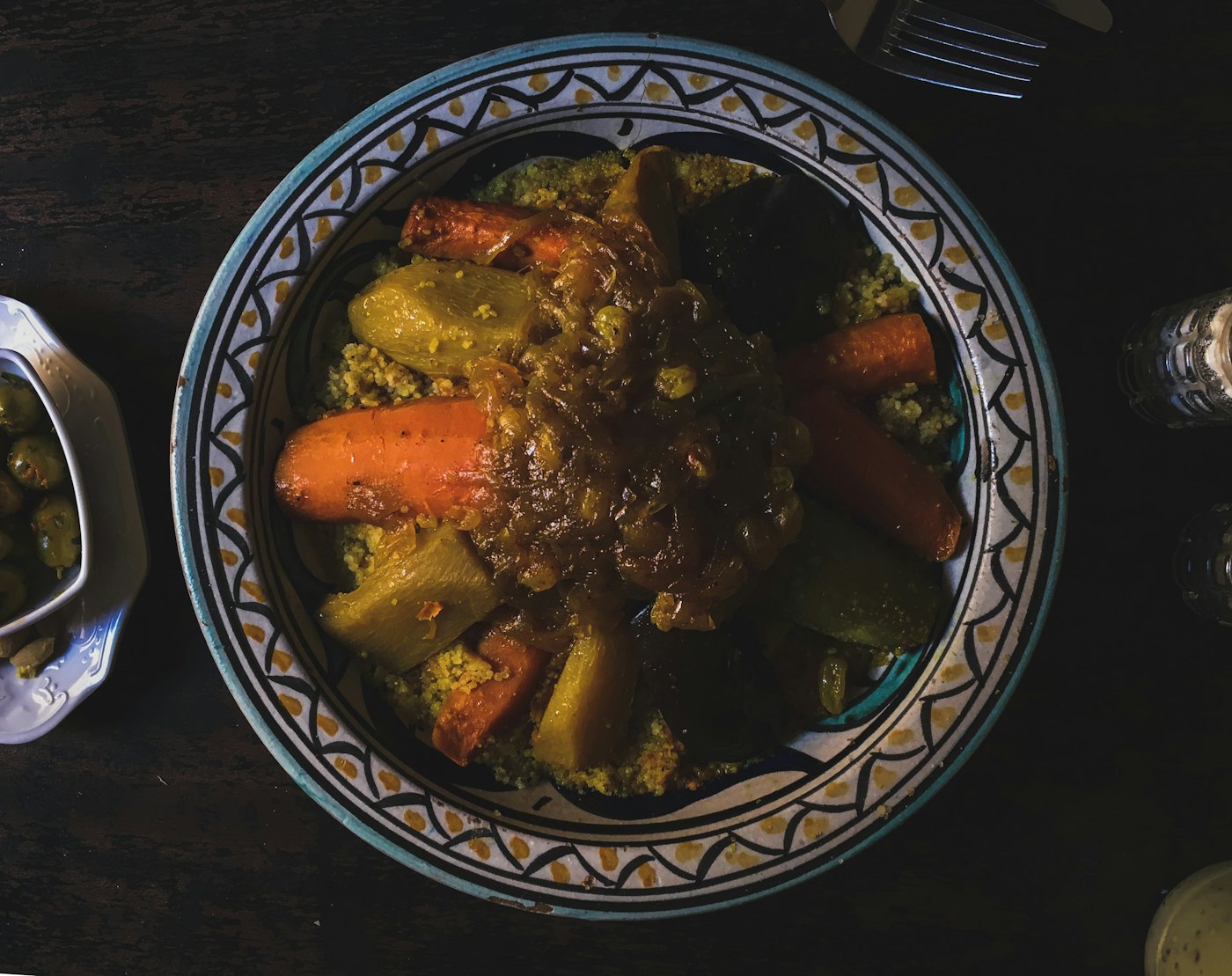 Plat de faïence orné garni de semoule, de carottes confites et de viande mijotée, photographié de haut dans un clair-obscur profond