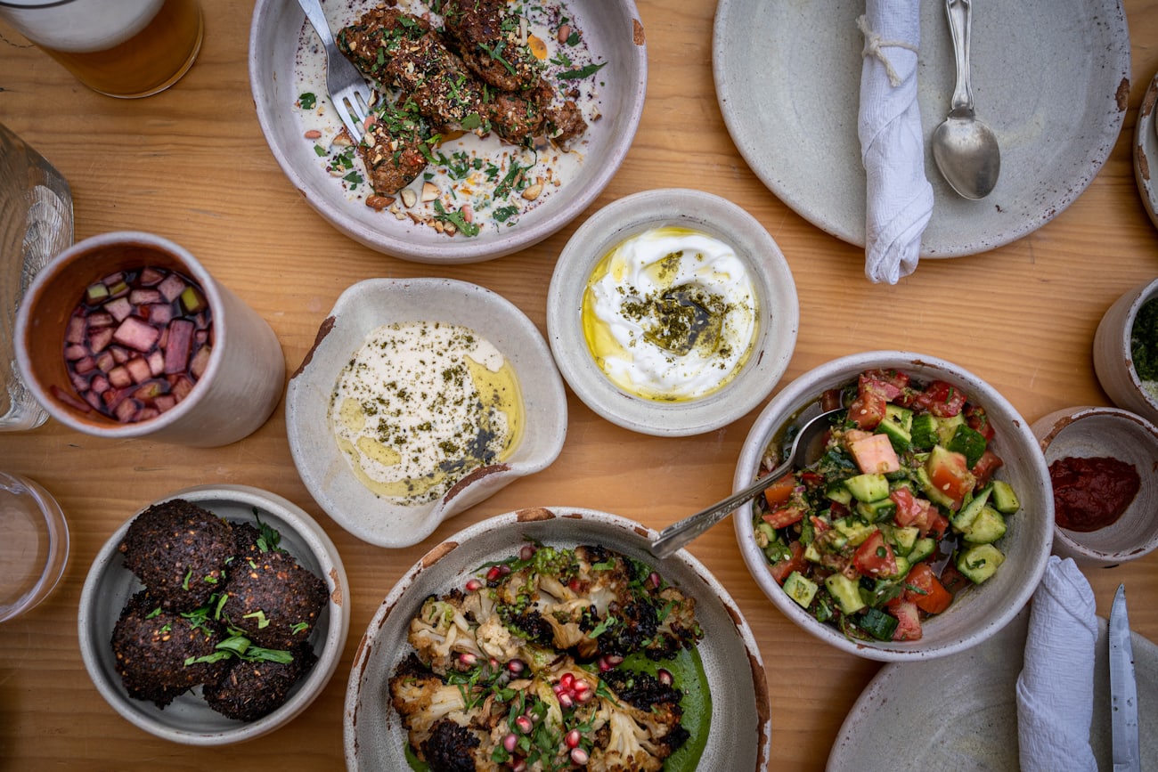 La table de mezze vue du dessus, sur bois clair : kefta d'aubergine au tahiné, houmous, labneh à l'huile d'olive, salade de tomates et concombre, falafels et chou-fleur rôti à la grenade
