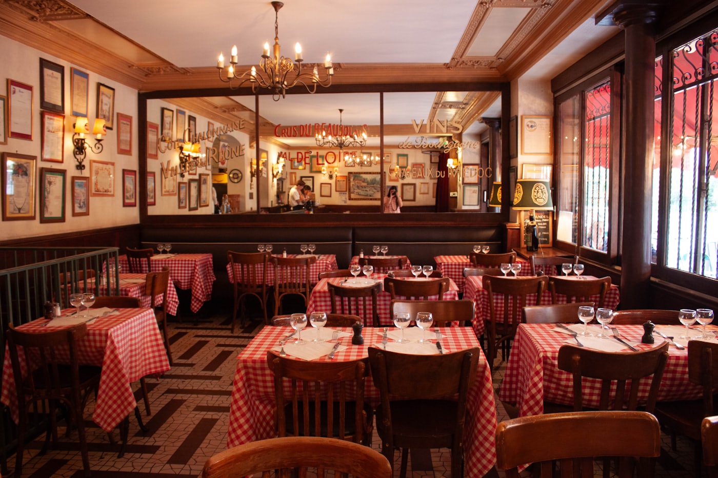La salle du bistrot : nappes vichy rouges, banquette, grand miroir peint aux noms des crus, lustre et sol en mosaïque.