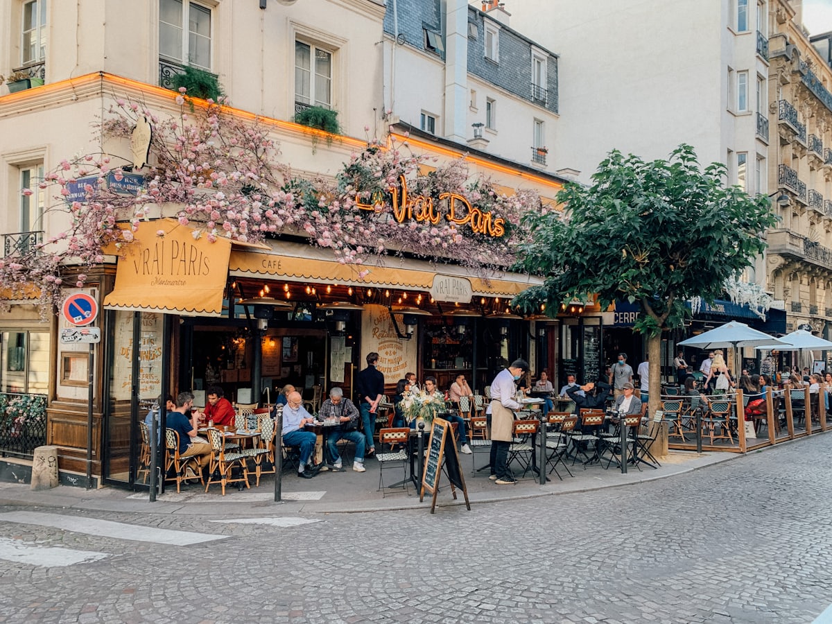 Façade fleurie et terrasse d'un café parisien : clients attablés au soleil, rue pavée bordée d'immeubles haussmanniens.