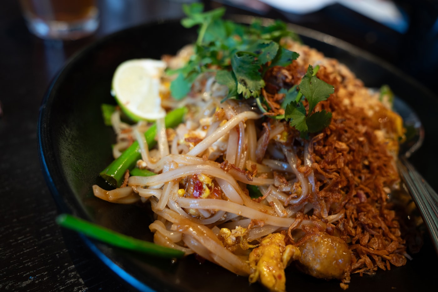 Pad thaï aux crevettes servi dans un bol noir, quartier de citron vert et coriandre fraîche, sur fond sombre.