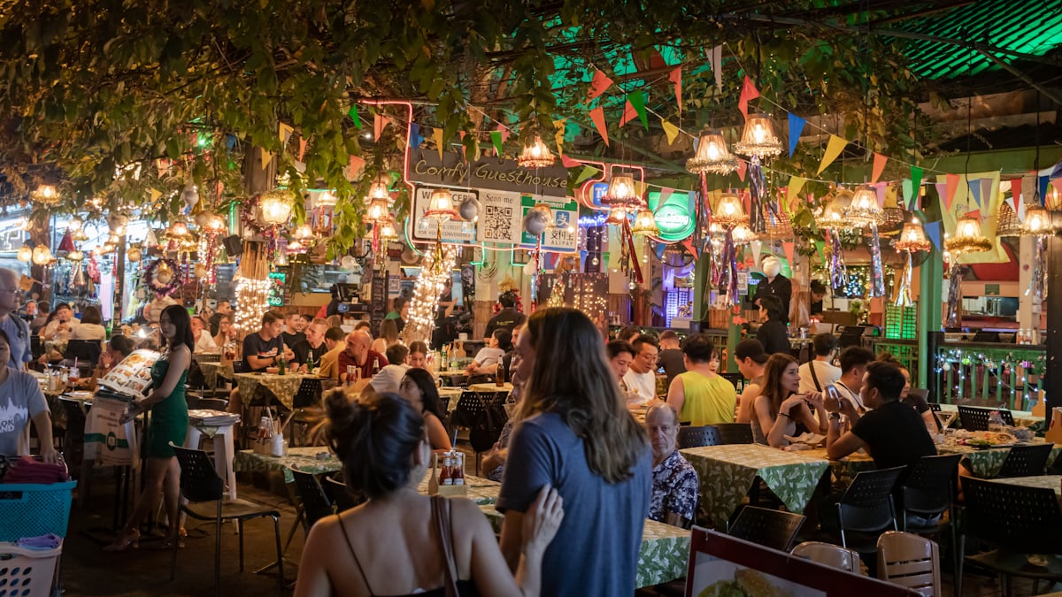 Terrasse de street food asiatique la nuit : guirlandes d'ampoules tendues au-dessus de tablées animées, verdure et lumière chaude.