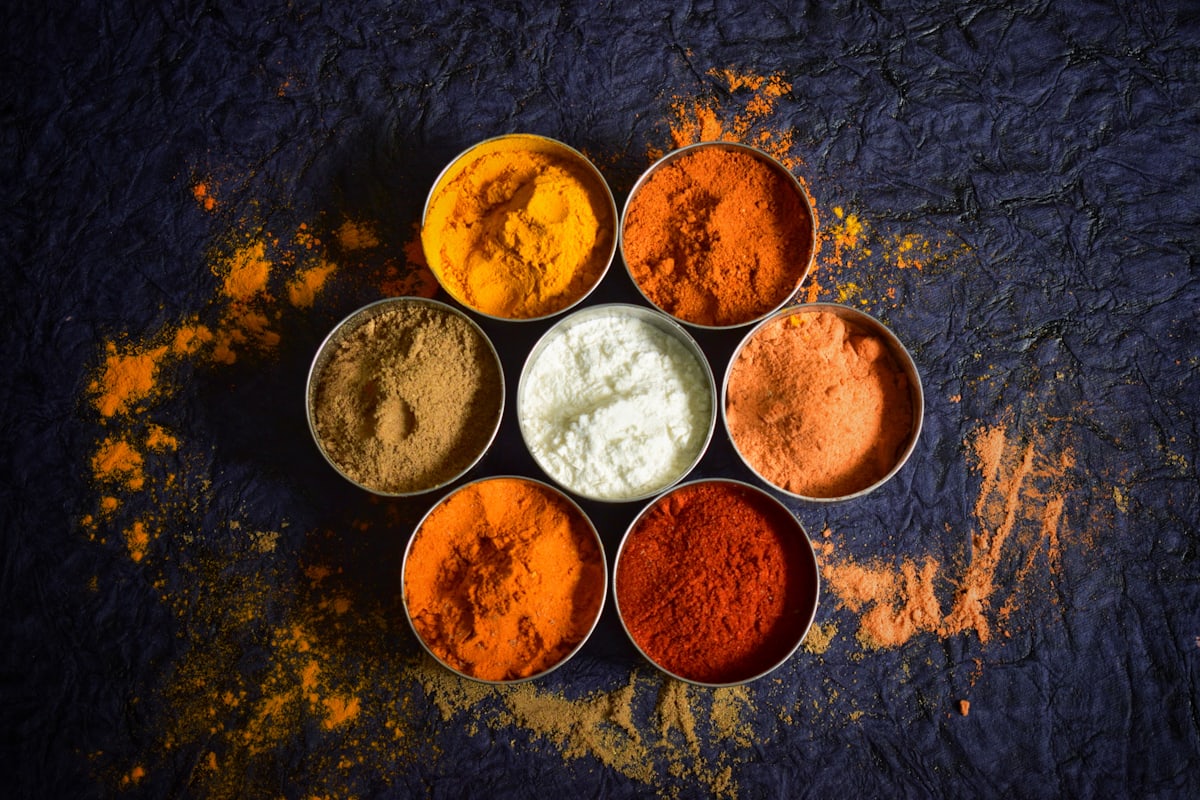 Sept épices en poudre — curcuma, paprika, cumin… — dans des coupelles rondes en inox sur fond noir, pigments renversés autour.