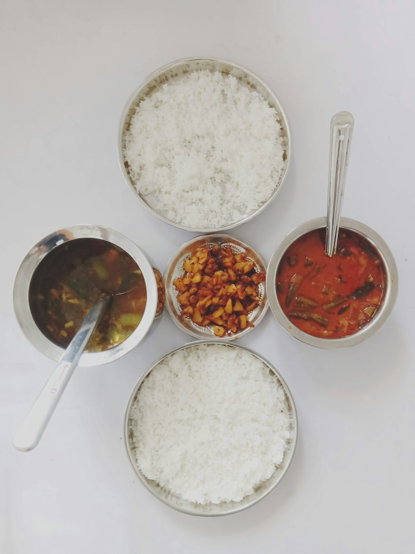La gamelle ouverte, vue du dessus : quatre boîtes rondes en inox disposées en cercle — riz basmati, curry sombre, curry rouge aux okras, condiments au centre.