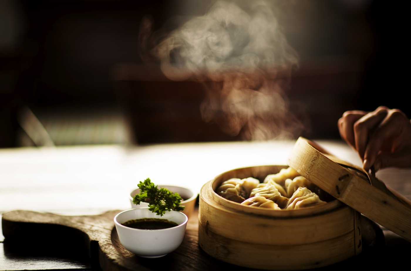 Une main soulève le couvercle d’un panier vapeur en bambou&nbsp;: la vapeur monte en pleine lumière au-dessus des dumplings pliés à la main.